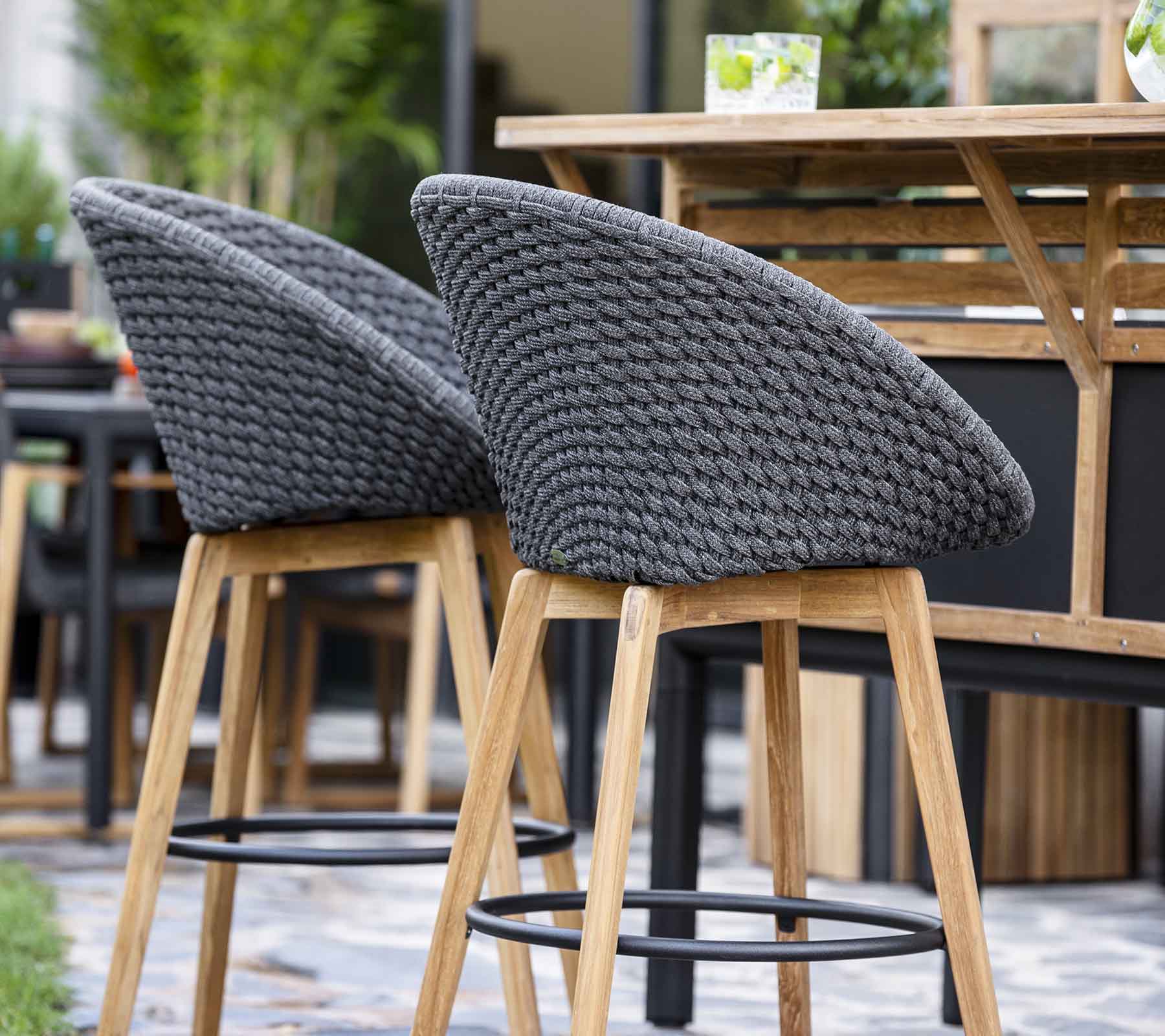 Stylish bar stools with woven gray backs and wooden legs, arranged near a wooden table in a contemporary setting.