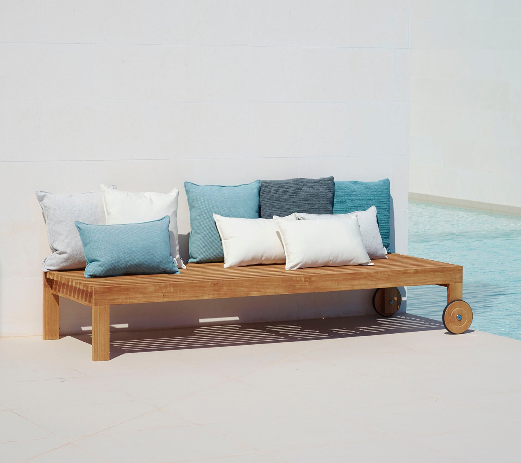 Wooden lounge sofa with a simple design, featuring cushions in shades of blue and white, positioned beside a pool.