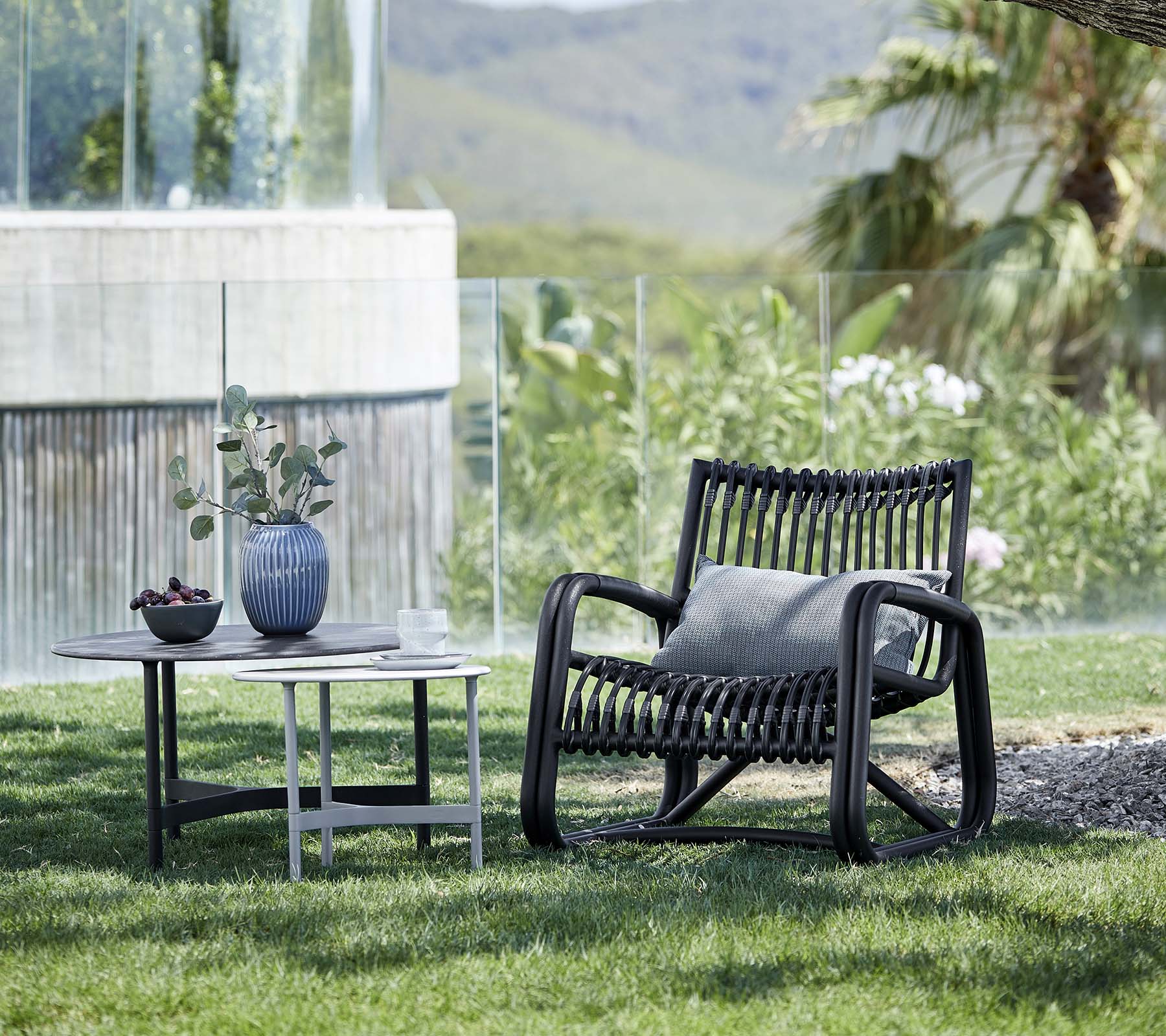 Contemporary lounge chair with a patterned design, accompanied by a side table holding a vase and potted plant, set in a natural landscape.