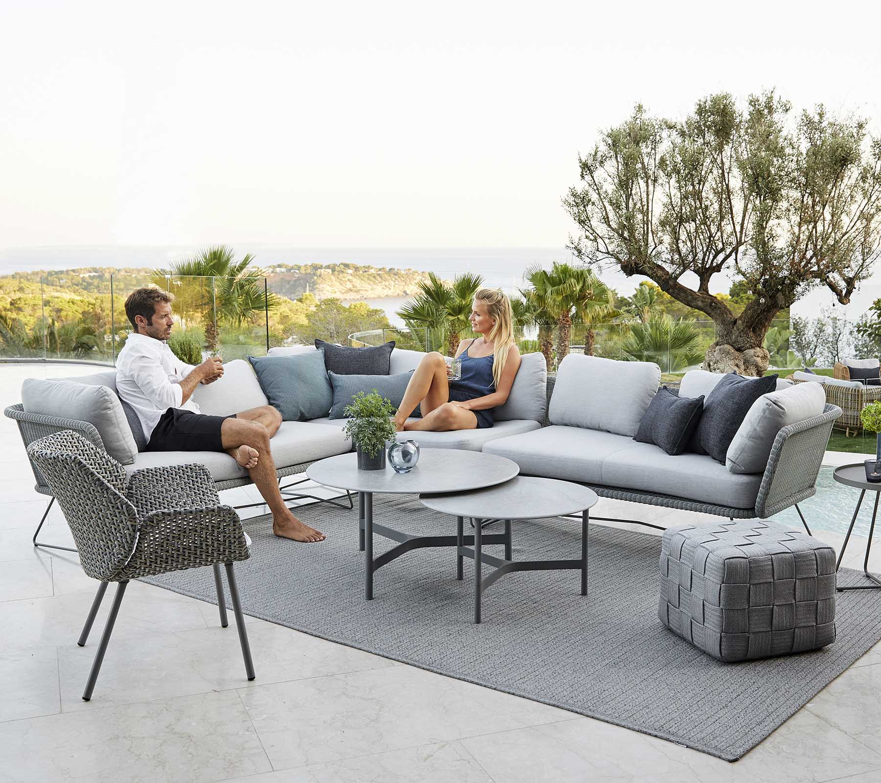 A modern outdoor seating area featuring a couch, armchair, and coffee table, with two people relaxing surrounded by greenery and a scenic view.