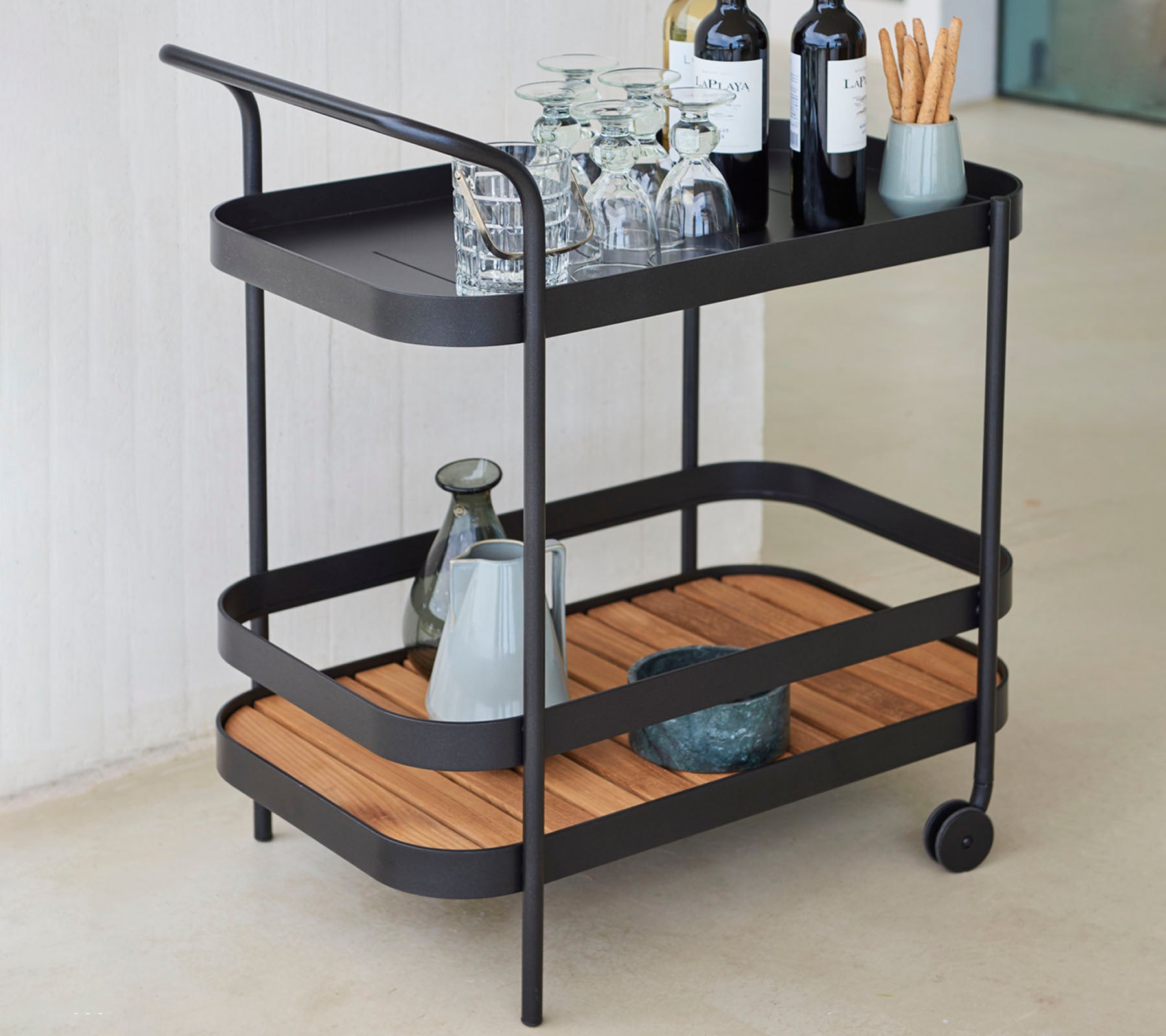 Stylish serving cart featuring two levels, glassware, wine bottles, and a vase, with a mix of metal and wooden design elements.