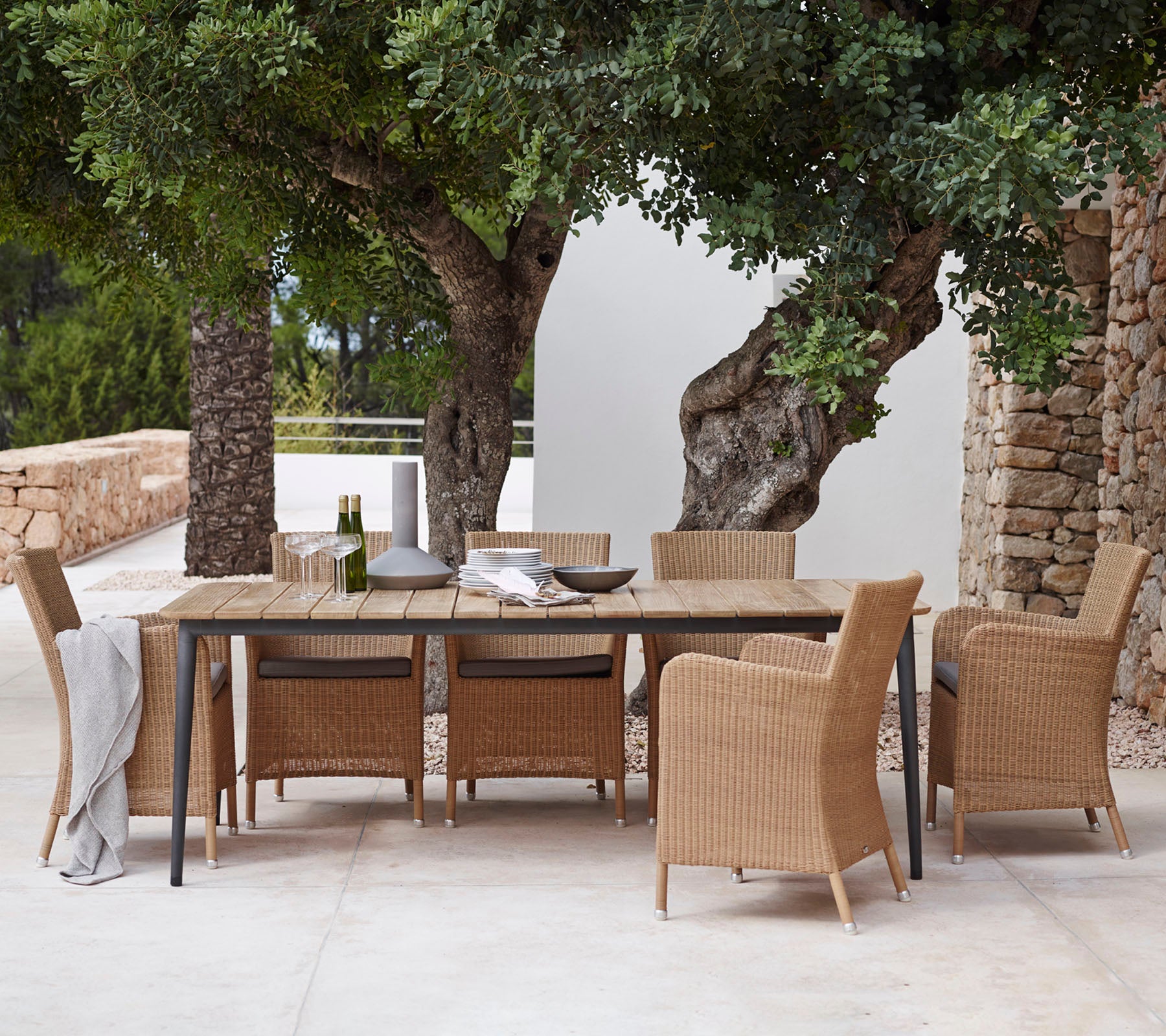 A stylish dining set featuring a long table and woven chairs, set beneath lush greenery and surrounded by natural stone walls.