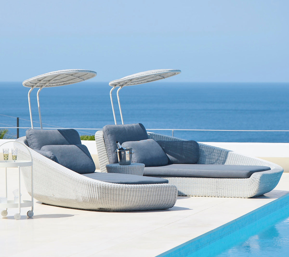 Stylish outdoor lounge chairs with canopies beside a serene pool, overlooking a tranquil sea. Perfect for relaxation and leisure.