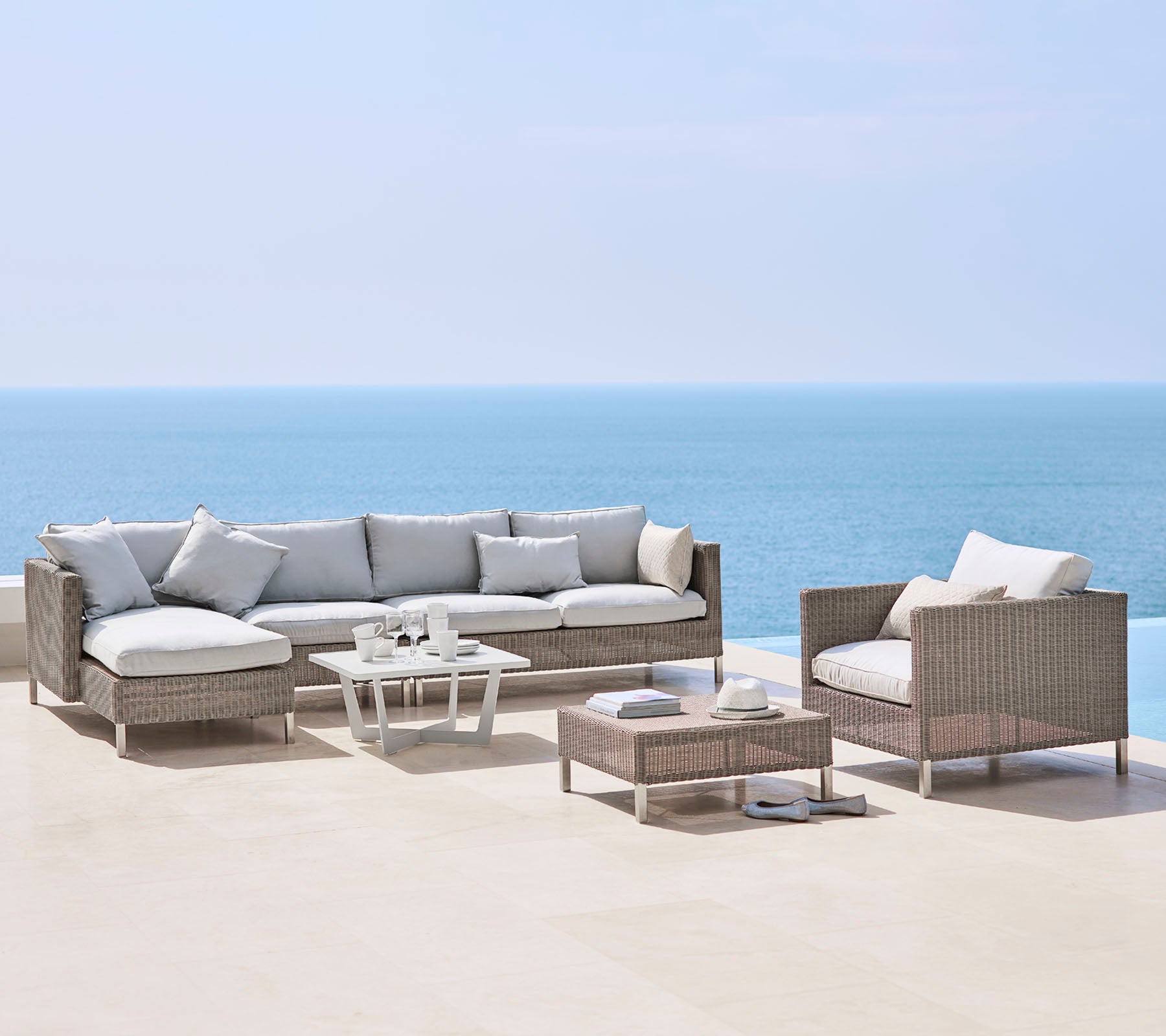 A stylish outdoor seating arrangement with a large sofa, an armchair, and a coffee table, all featuring light cushions and woven material, by the sea.