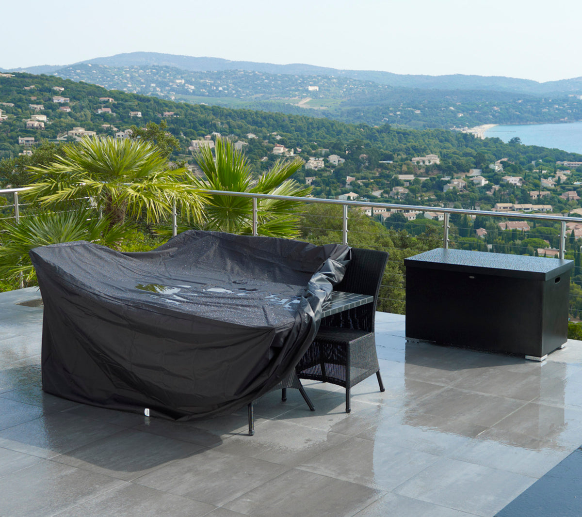Outdoor seating area covered with a black protective cover, accompanied by a matching storage box and a scenic view of hills and water.