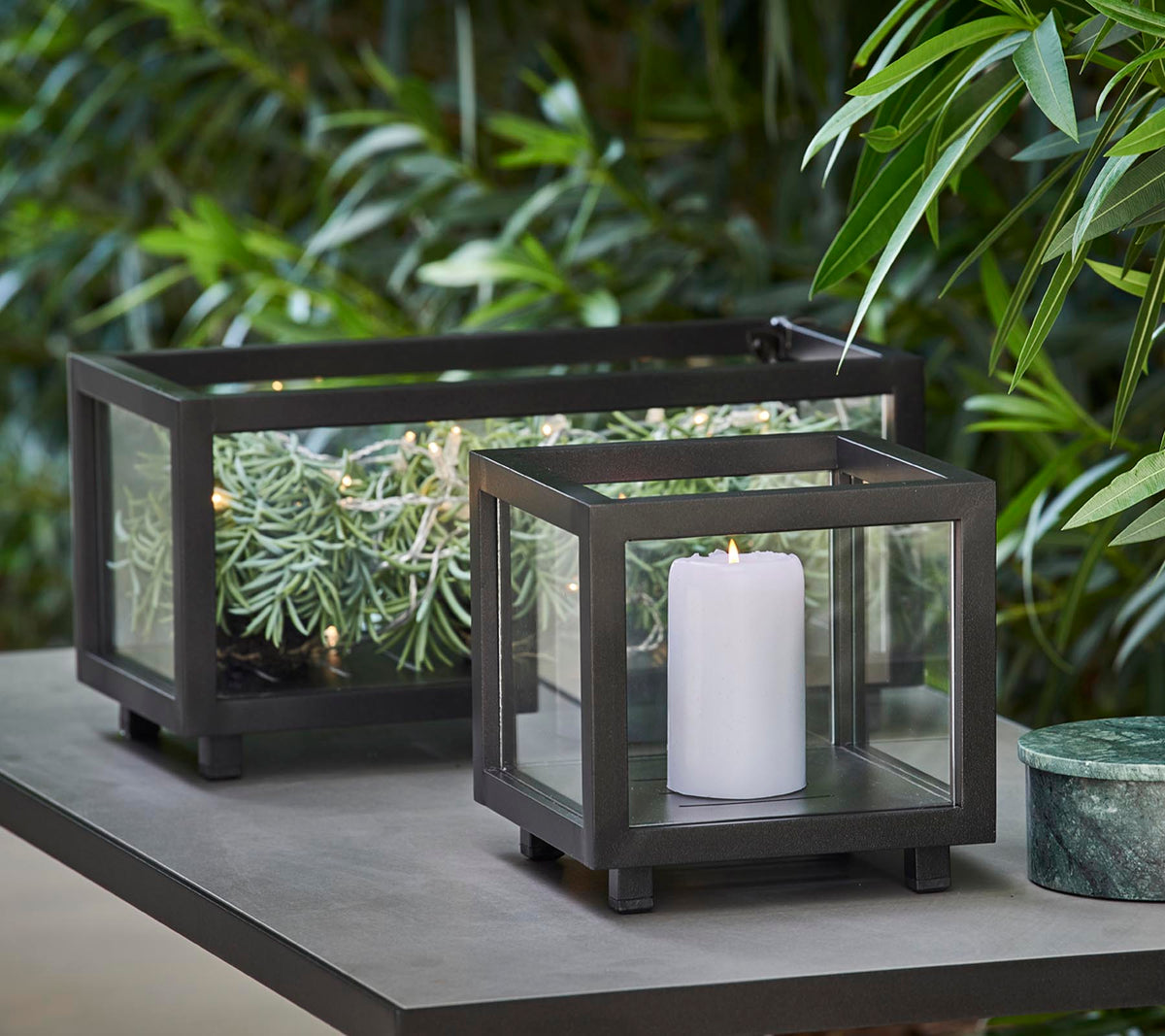 Two stylish lanterns with glass panels, one holding a white candle and the other featuring greenery, placed on a table amid lush foliage.