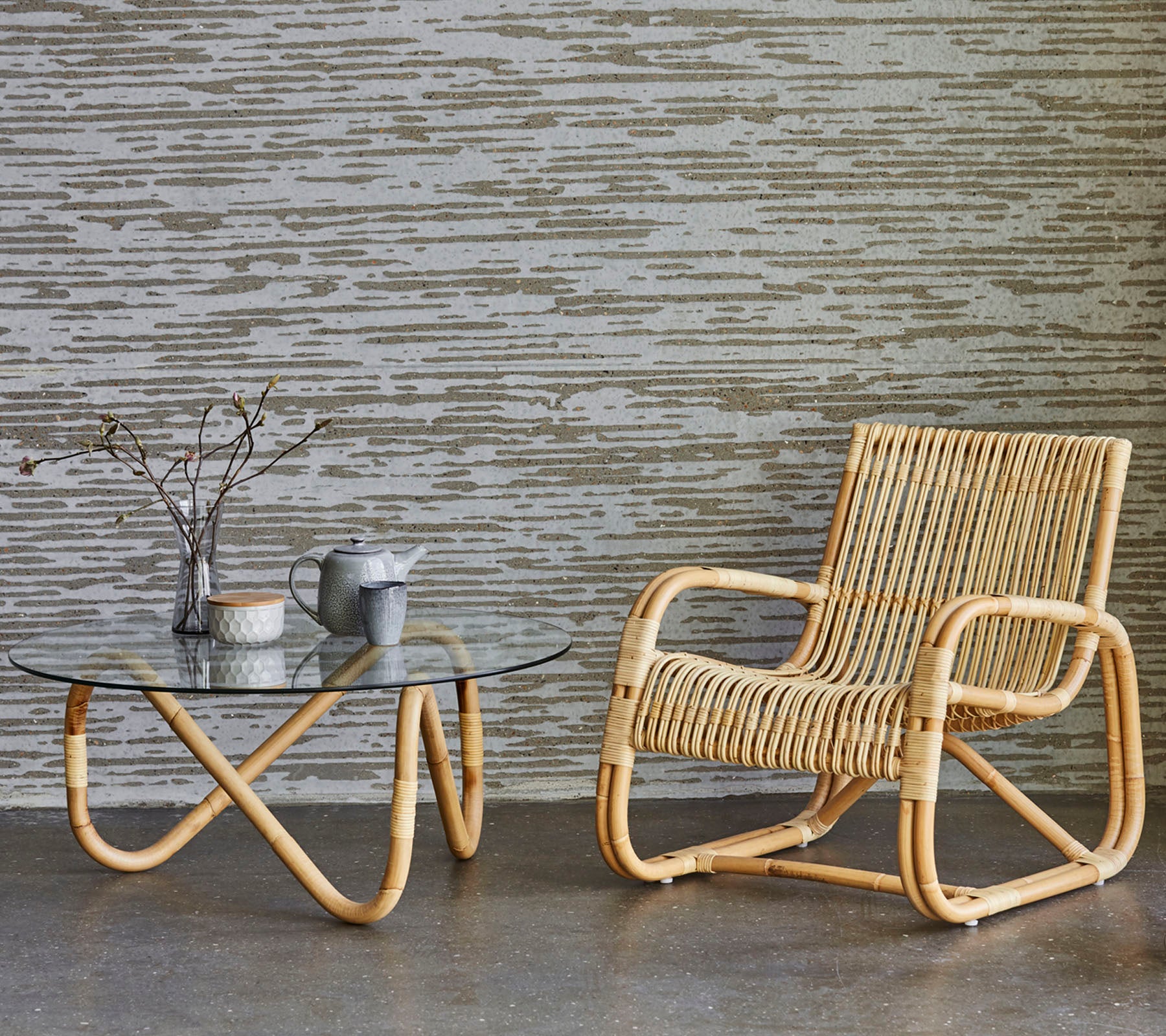 Rattan chair and glass-topped table with decorative items, set against a textured wall. A minimalist and modern decor style.
