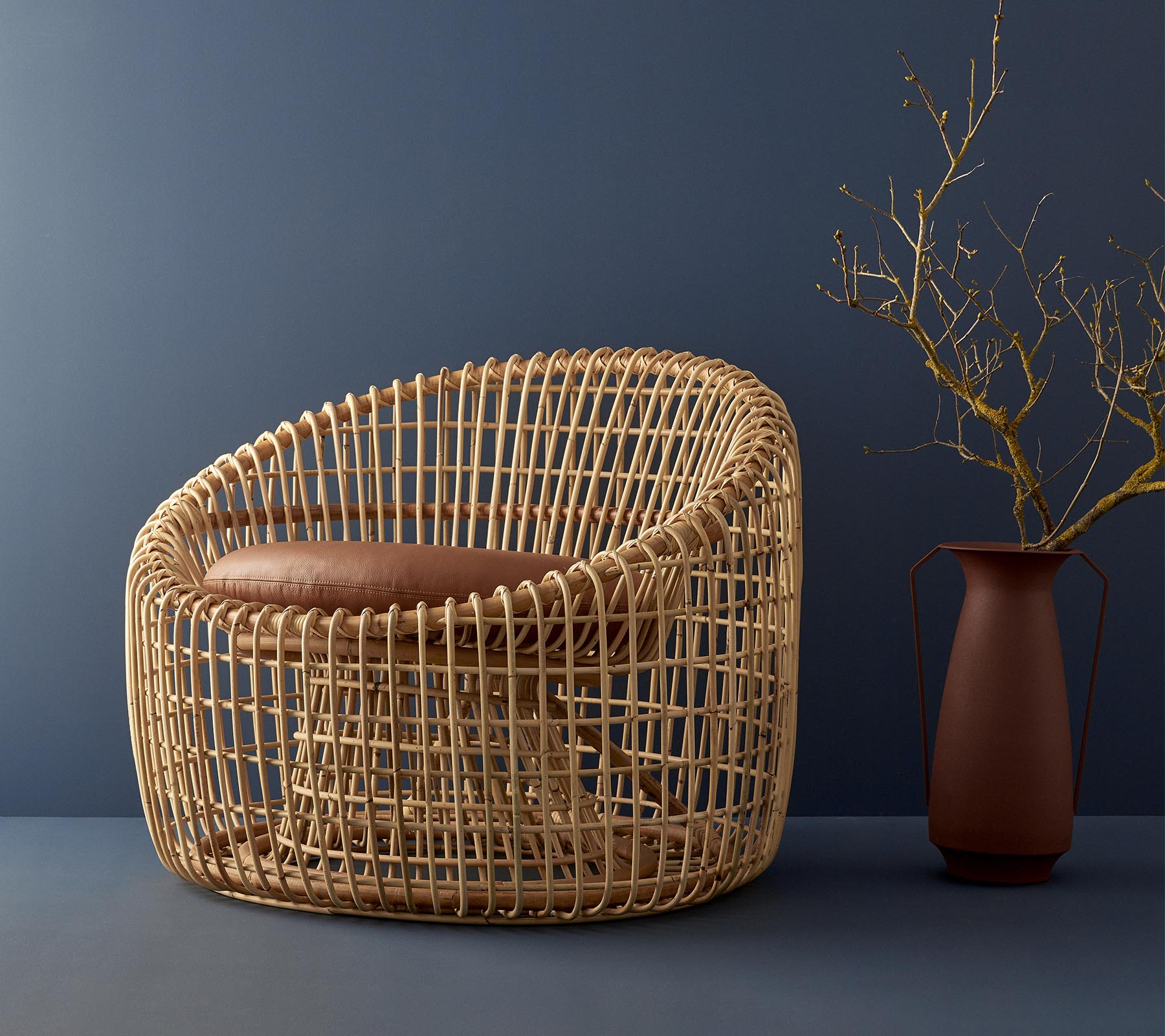 Modern wicker chair with a rounded design, featuring a cushioned seat and complemented by a simple vase with a twig.