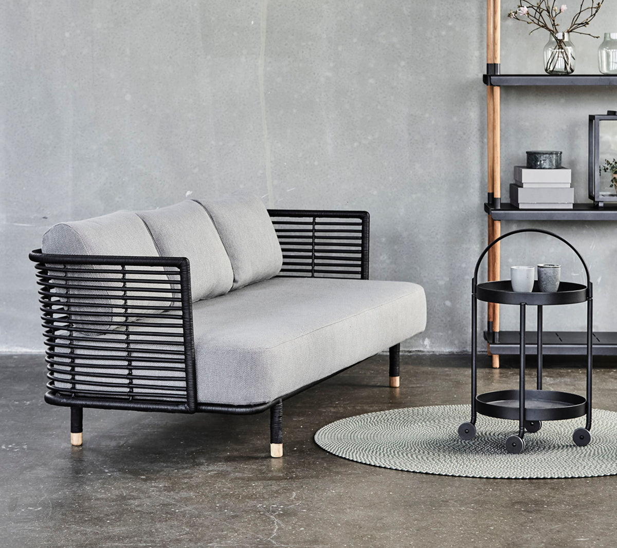 Sleek gray sofa with black metal accents and cushions, paired with a round side table on a textured rug against a neutral background.
