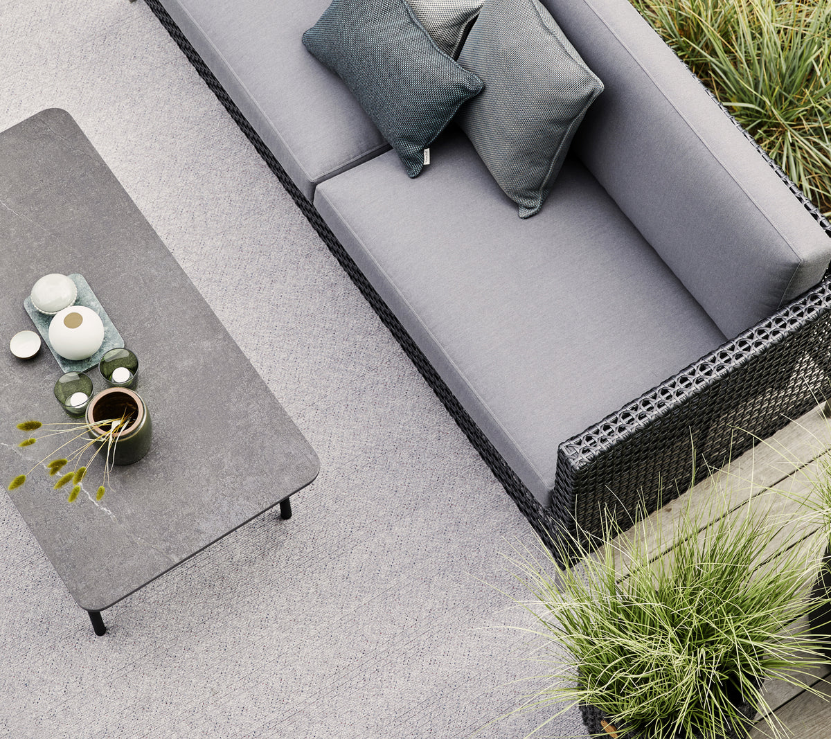 A gray sofa with two pillows sits near a minimalist table adorned with ceramics and a small plant, surrounded by greenery.