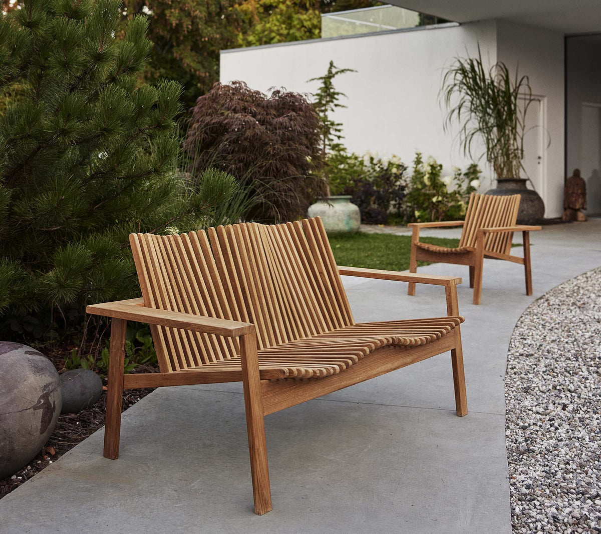 Two wooden Amaze lounge chairs and a matching sofa feature a slatted design, surrounded by greenery and decorative elements.