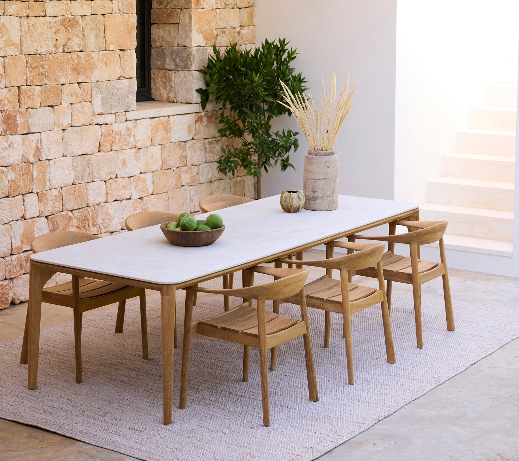 Modern dining setup with a white table and wooden chairs against a stone wall.