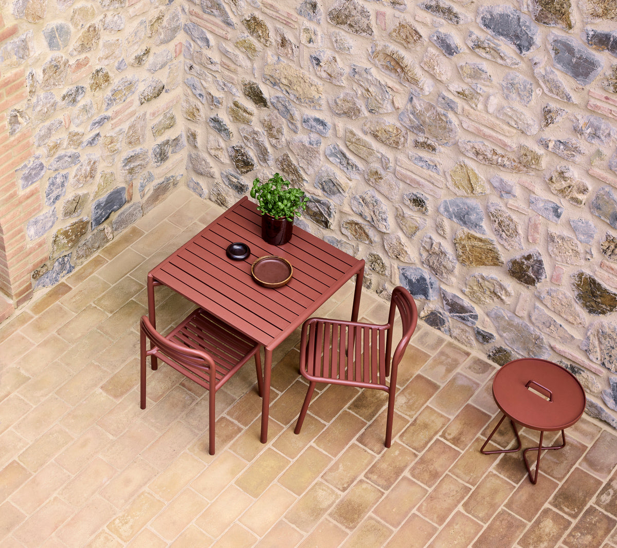 A red cafe table set with two chairs, featuring a small plant and a plate, against a stone wall backdrop.