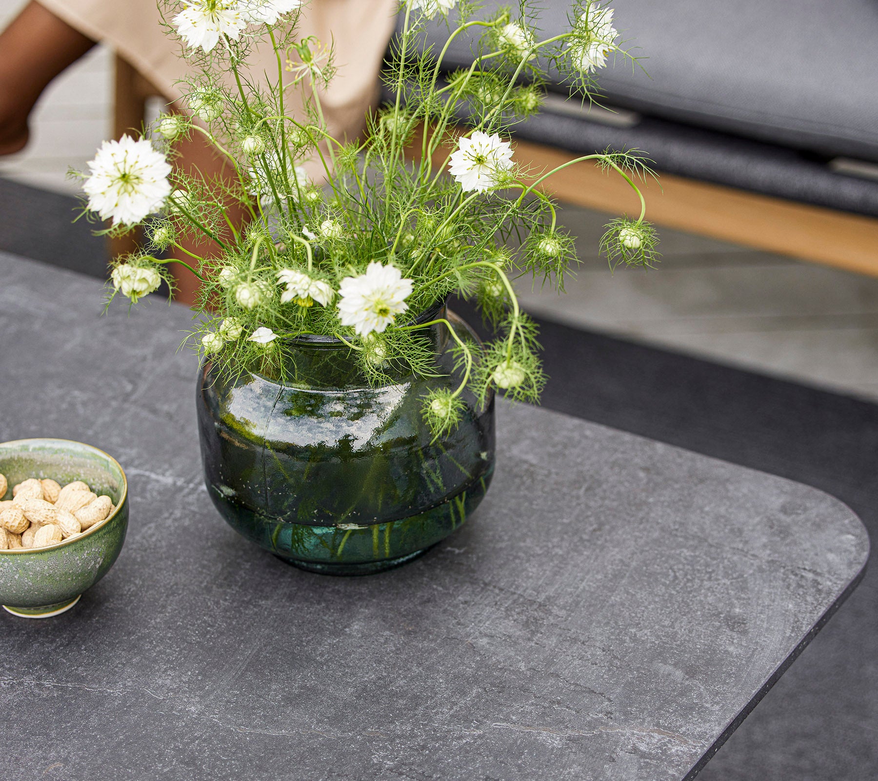 A modern tabletop featuring a round glass vase with white flowers and a small bowl of nuts. The surface is sleek and textured.
