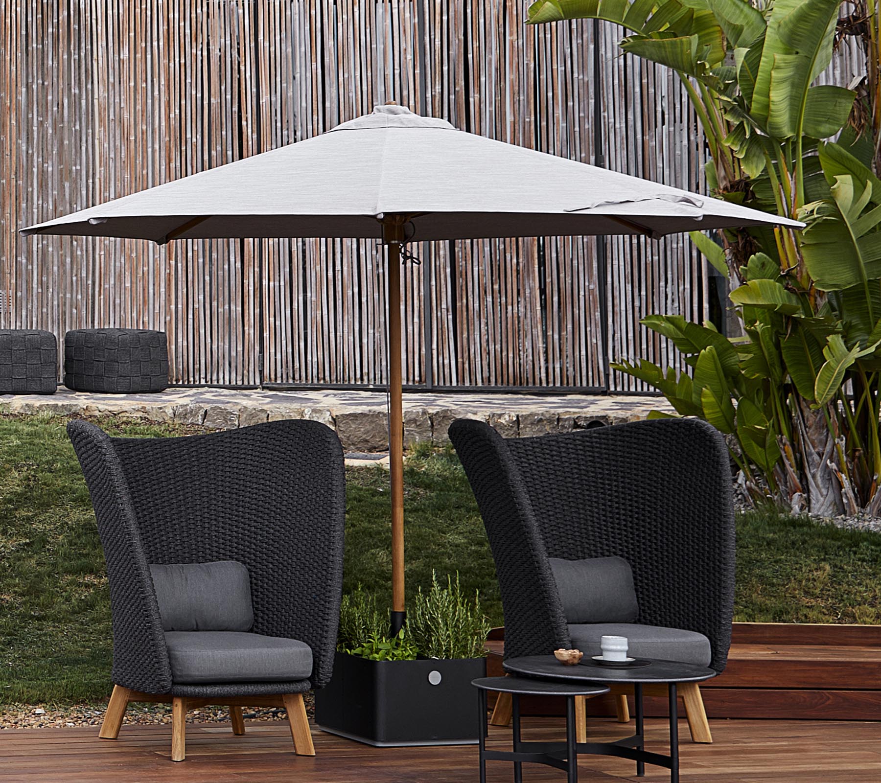 Two stylish woven chairs with cushions sit under a large umbrella, accompanied by a small table, set against a lush green backdrop.