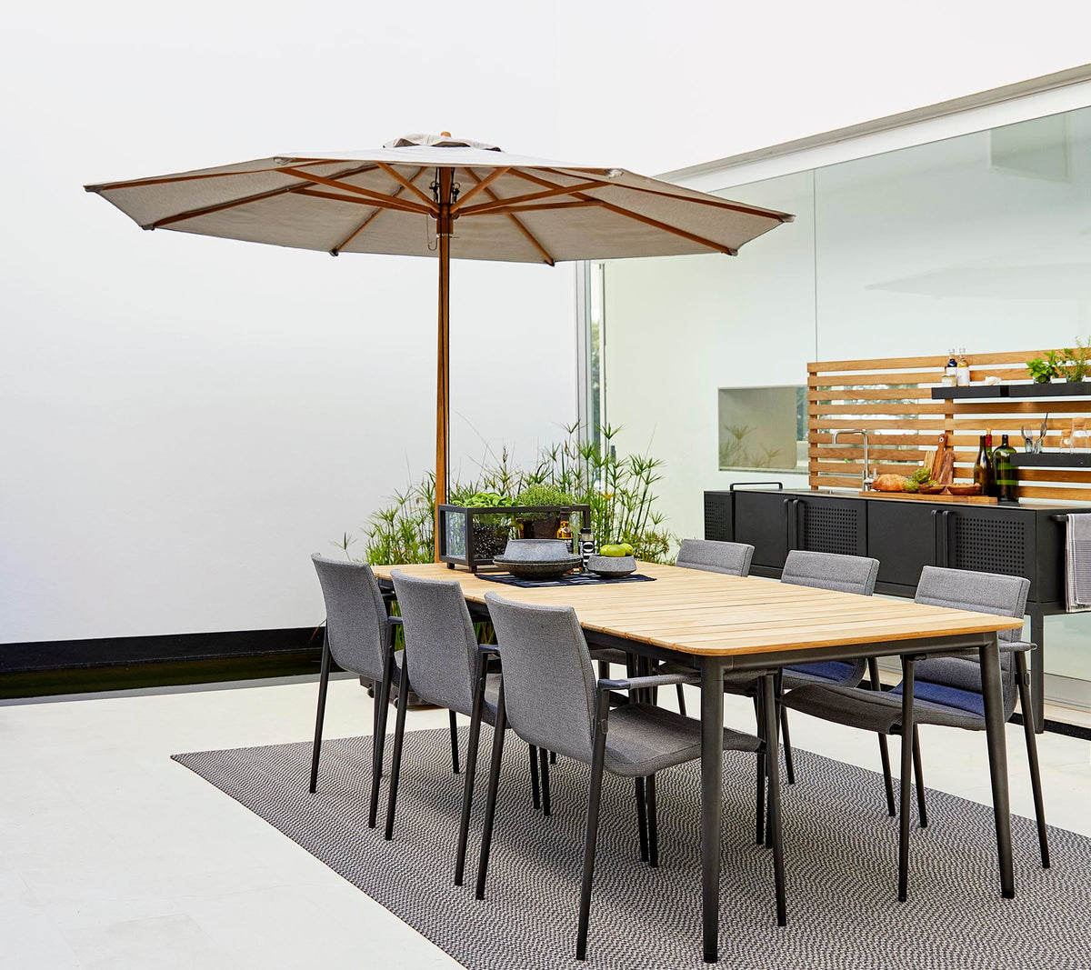 A classic dining set featuring a wooden table with an umbrella and gray chairs, surrounded by decorative plants on a stylish rug.