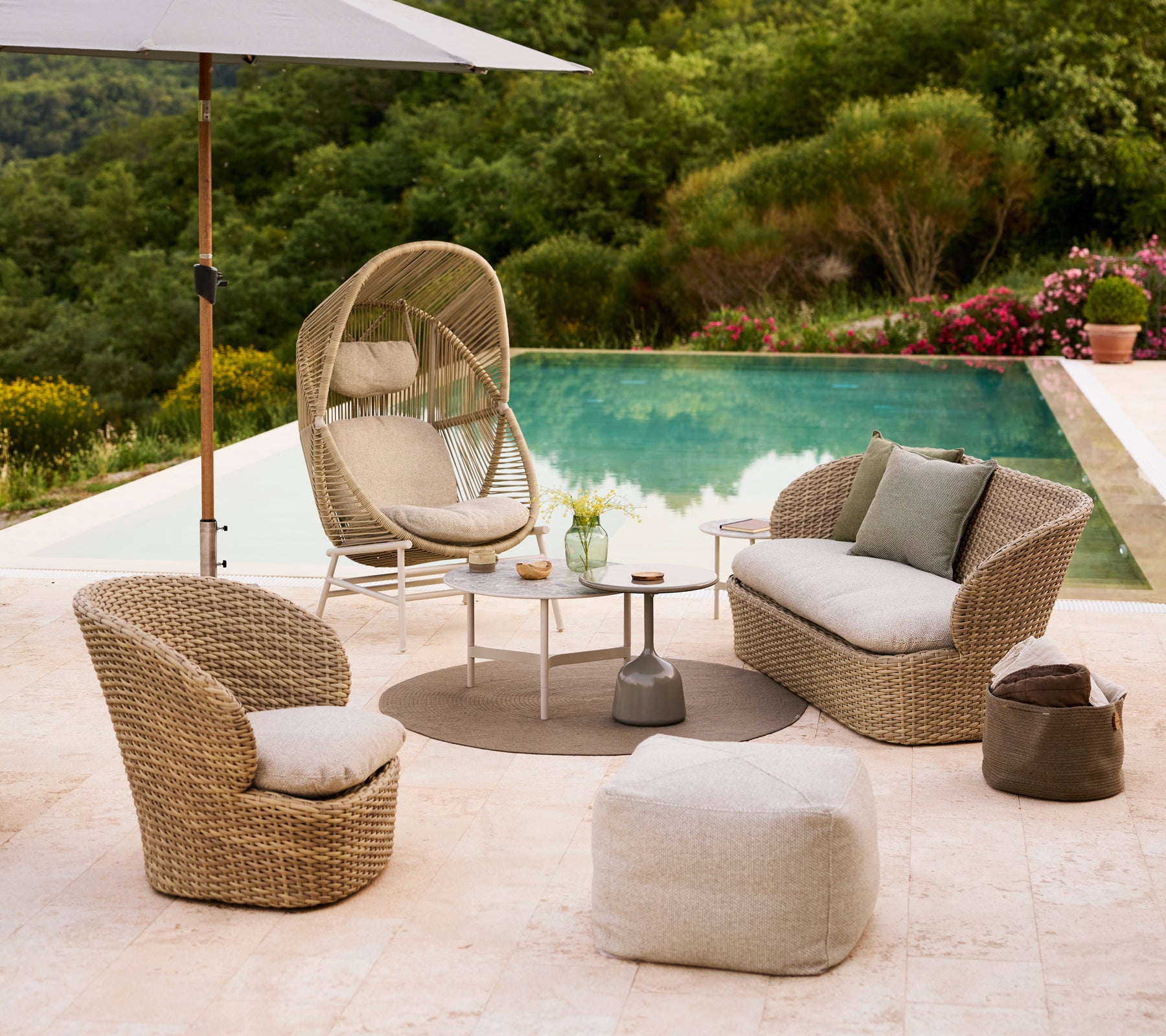 Coastal lounge setup featuring a Hive chair, two curved chairs, a Divine footstool, and a small table by a pool surrounded by greenery.