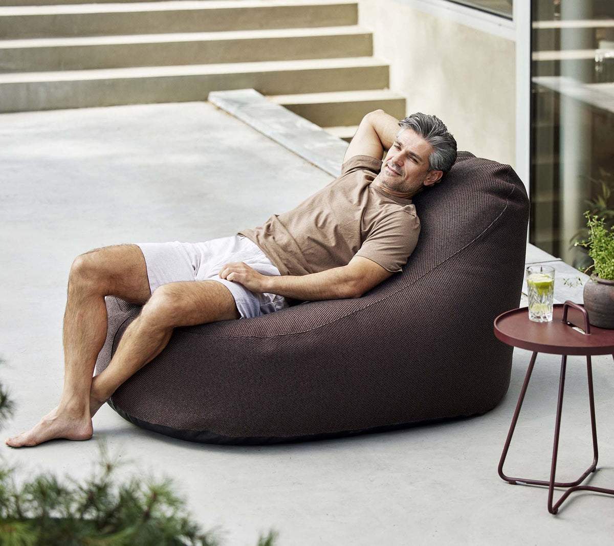 A man relaxes in a cozy bean bag chair, enjoying a drink beside a small table, surrounded by greenery.