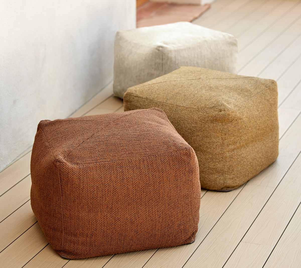 Three textured poufs in earthy tones of brown, beige, and tan, displayed on a wooden surface. Ideal for versatile seating and decor.