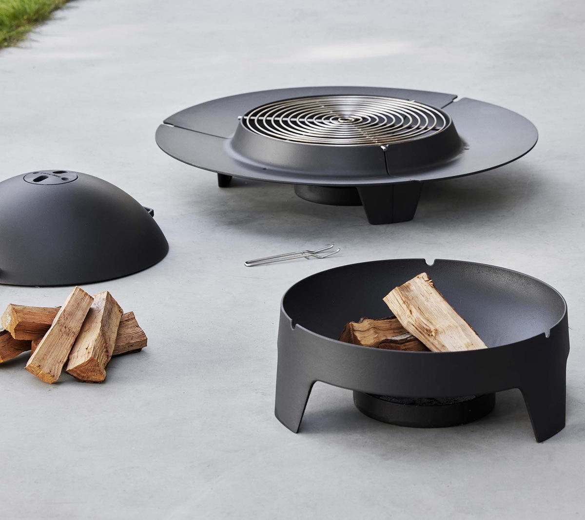 A sleek black fire pit with a grilling grate, a protective cover, and a base, accompanied by some stacked firewood.