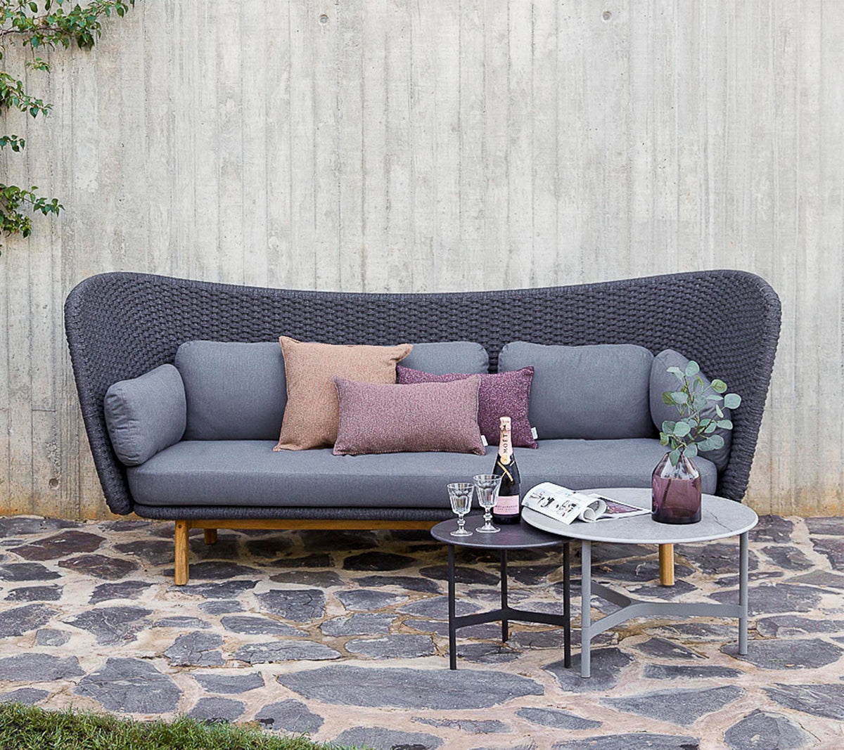 A modern sofa in grey with decorative pillows, accompanied by a small table with drinks and a vase, set against a textured wall.