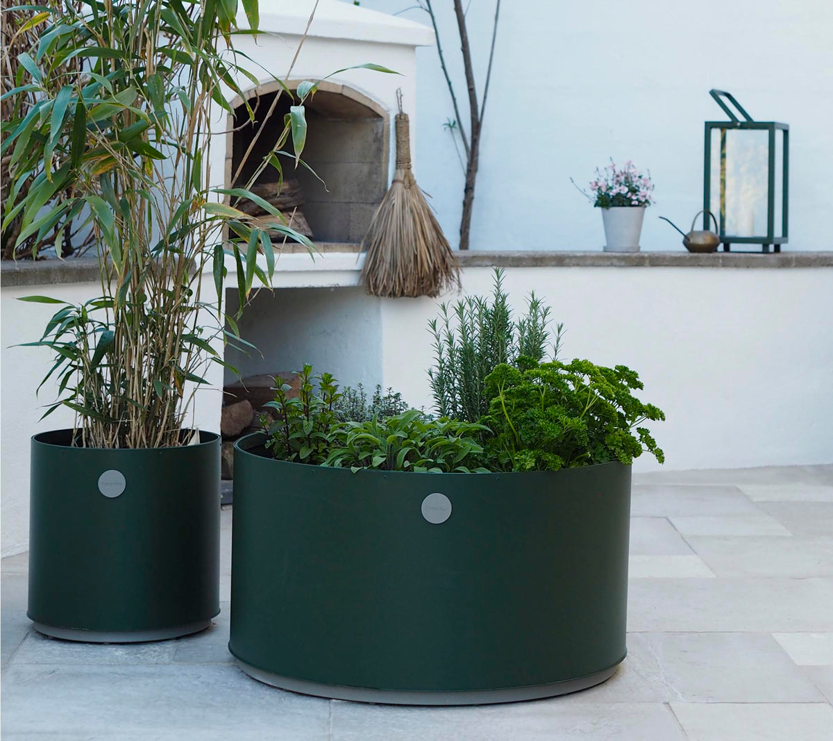 Two round planters in dark green, one featuring tall plants and the other filled with various herbs, sit on a light stone surface.
