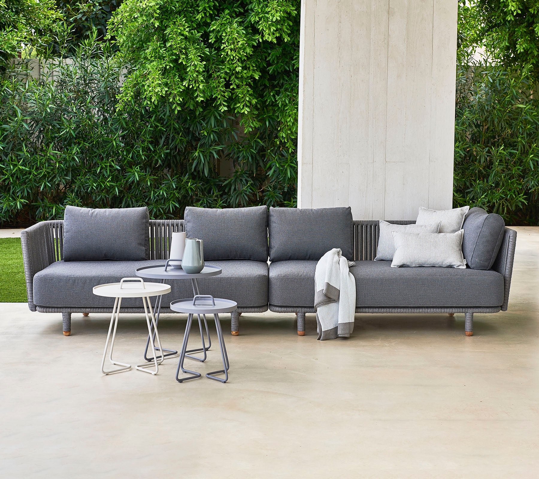 Modern gray sectional sofa with pillows, accompanied by two stylish side tables, set against a backdrop of lush greenery.