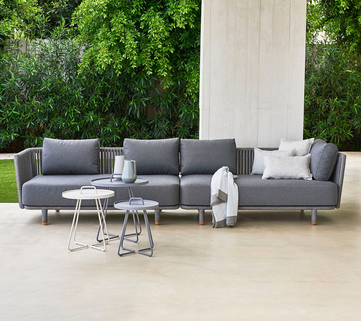 Modern gray sectional sofa with pillows, accompanied by two stylish side tables, set against a backdrop of lush greenery.