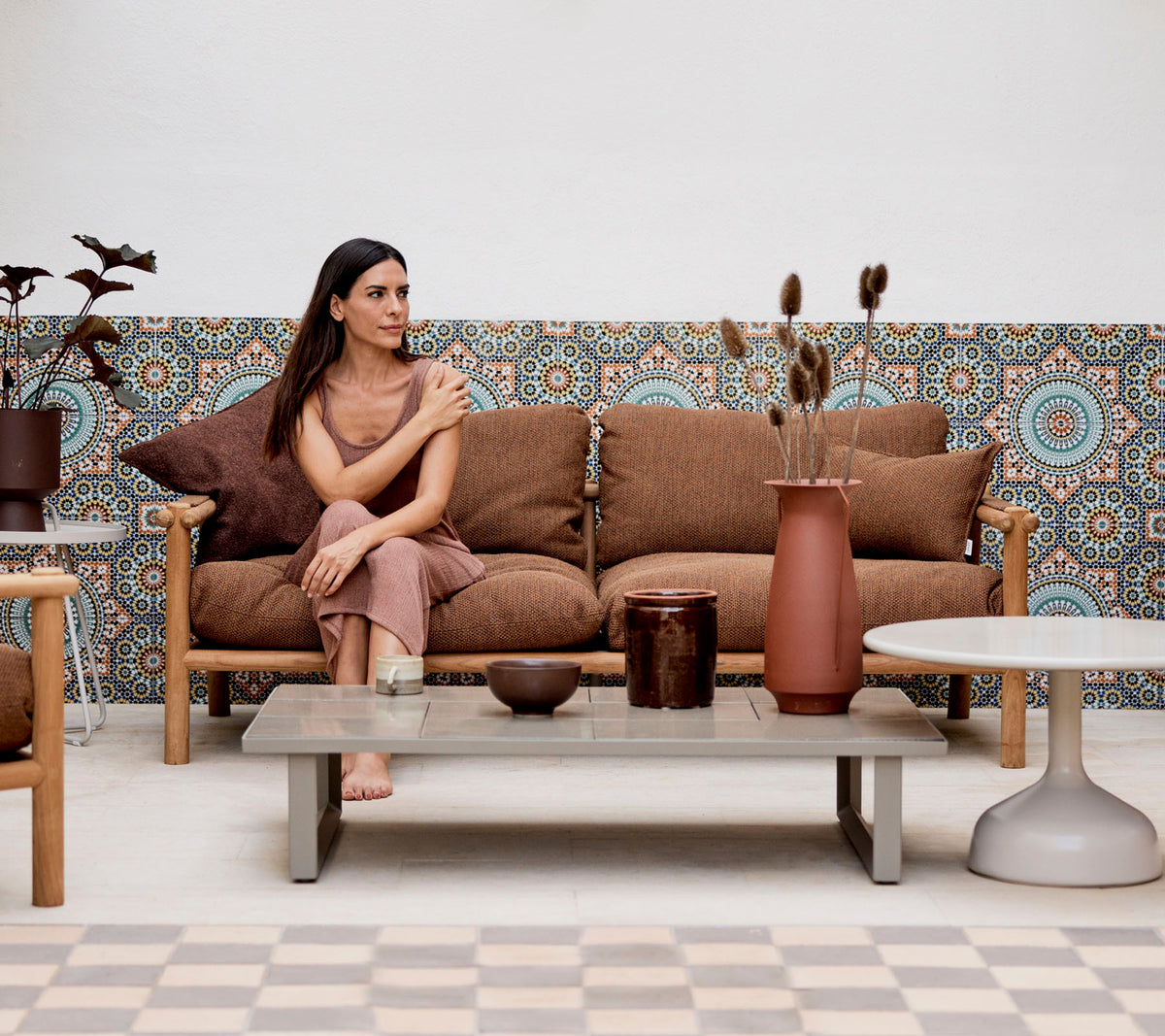 A woman seated on a couch with decorative pillows, surrounded by vases and plants, against a patterned background.