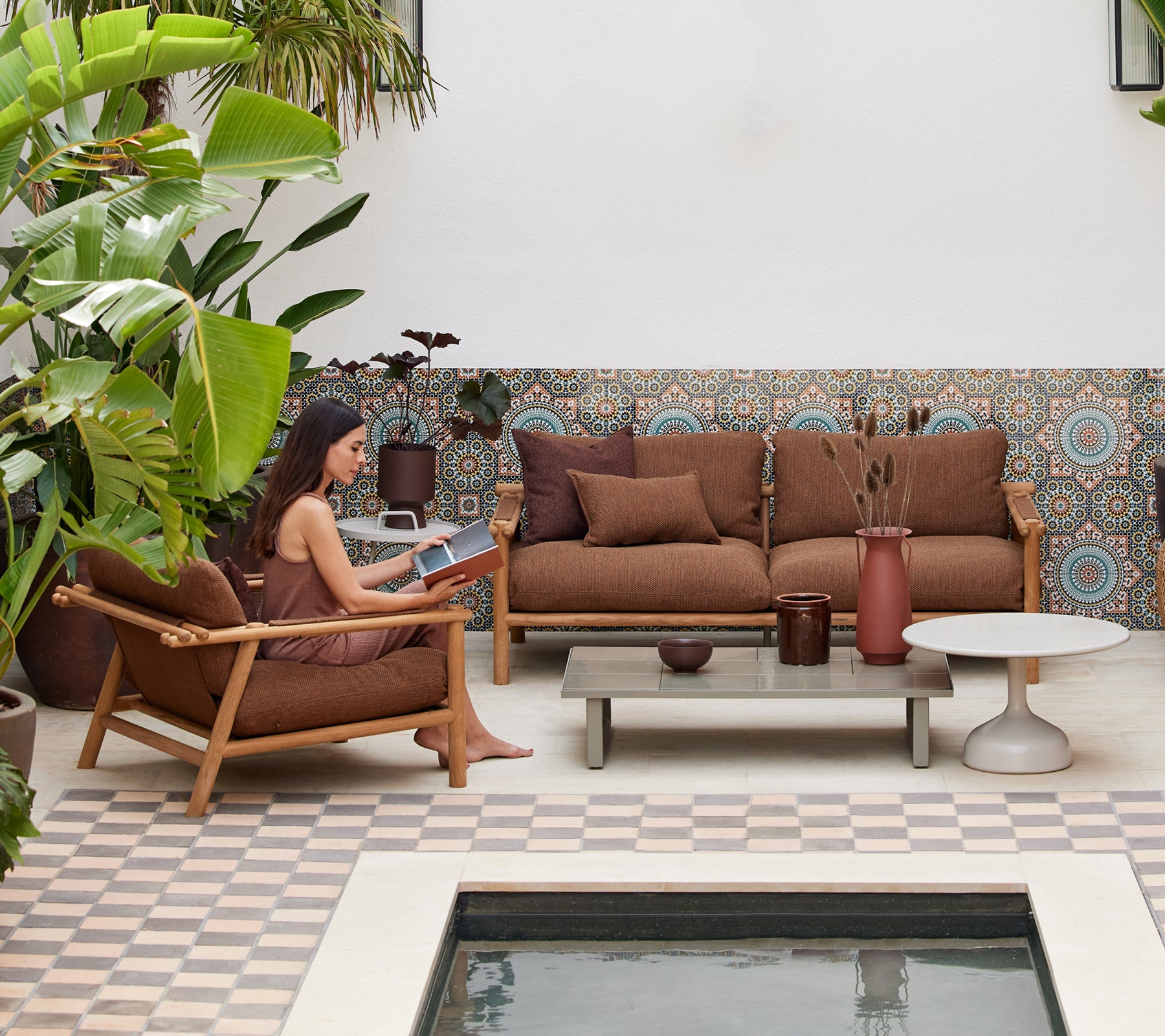 A person reading a book in a cozy lounge area featuring teak furniture, cushions, a coffee table, and surrounding greenery.