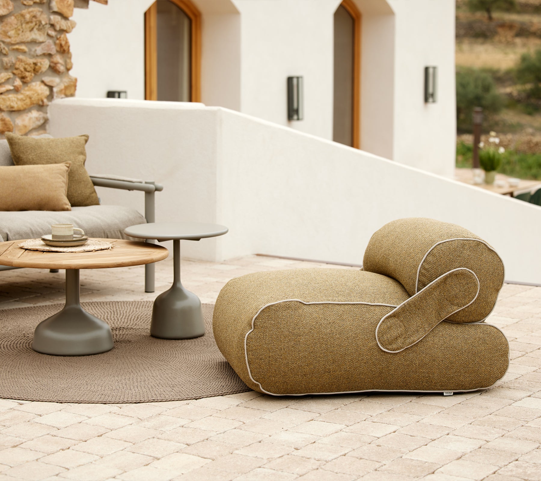 Stylish lounge chair with a unique shape, accompanied by a round table and decorative cushions, set in a tranquil environment.