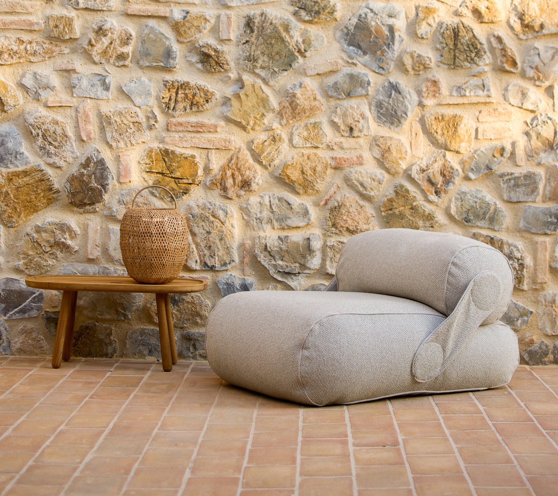 A stylish lounge chair next to a wooden coffee table and a decorative vase, set against a rustic stone wall.