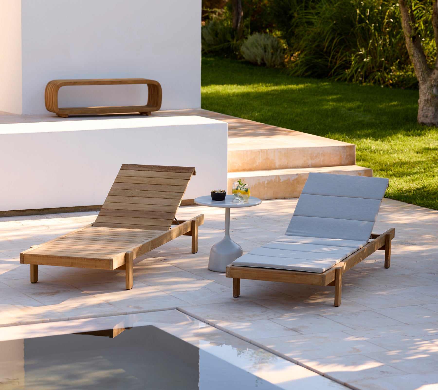 Two modern wooden loungers with grey cushions and a white side table in an outdoor setting.